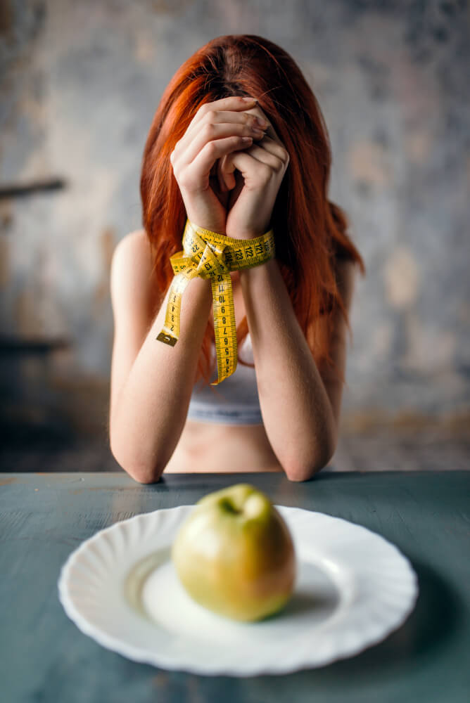 Exhausted young woman with hand tied with measuring tape, showing weakness and fatigue caused by anorexia nervosa and malnutrition.